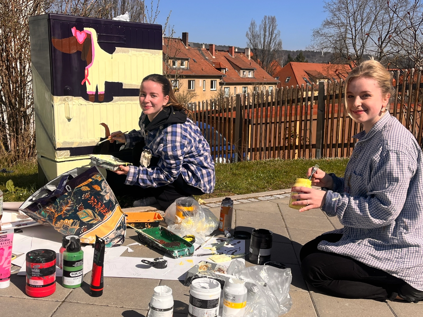 Schülerinnen verschönern einen Stromkasten mit Graffiti. 